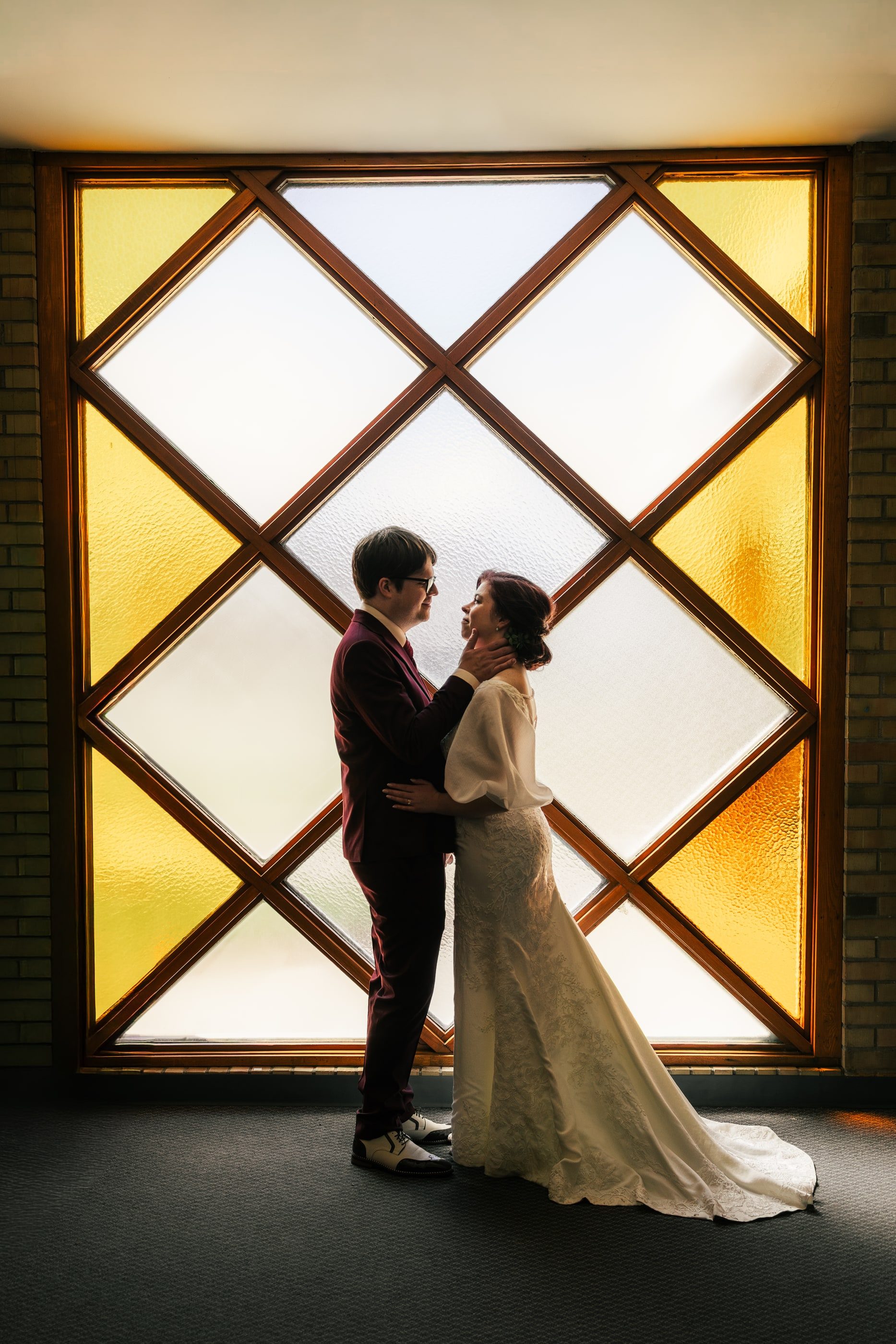 A couple in a burgundy suit and an ivory gown hold each other in front of a tall diamond-paned amber window.