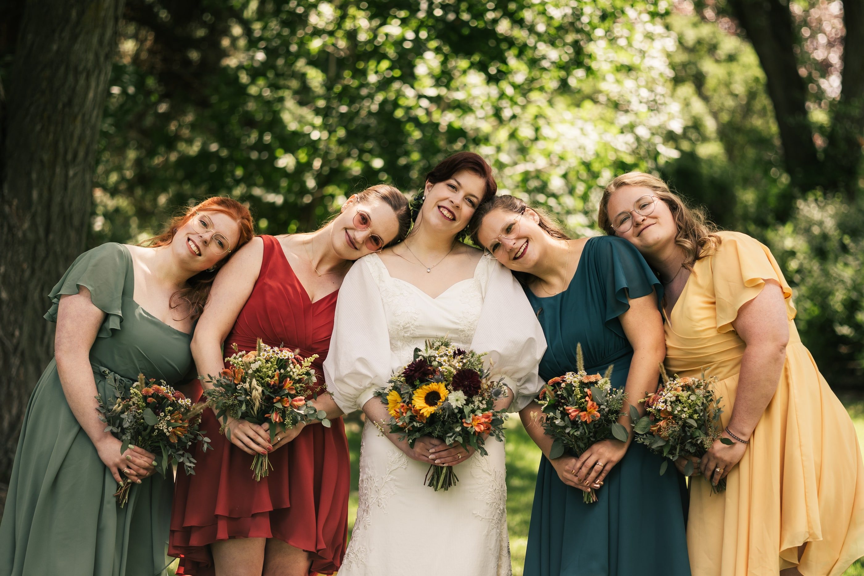 A bride leans in with her bridesmaids in green, red, teal and yellow dresses, all holding sunflower bouquets.