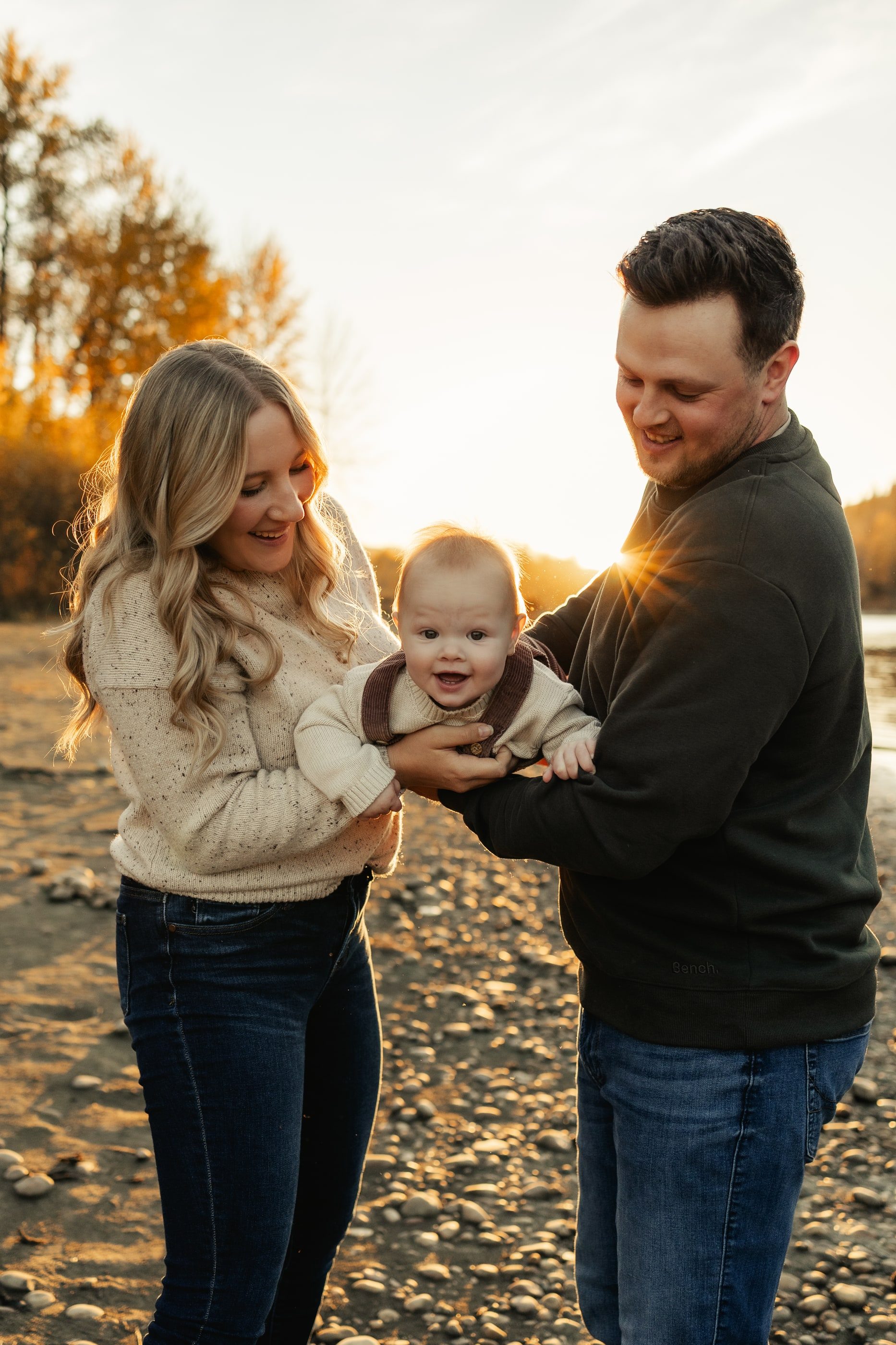 Parents laugh with their baby on a rocky riverbank in golden evening light.