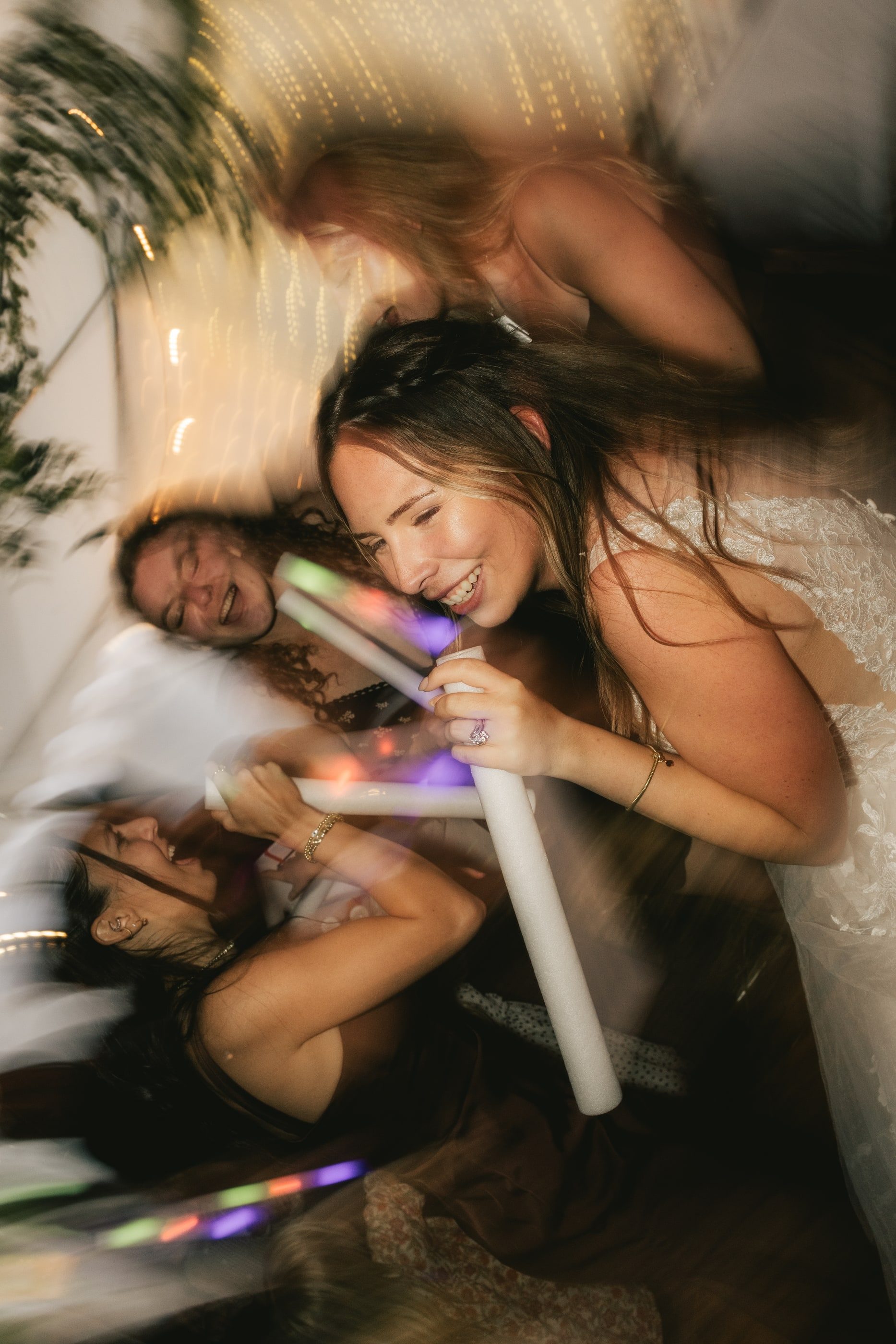 A bride and her friends dance and laugh with glowing light sticks on a crowded dance floor.