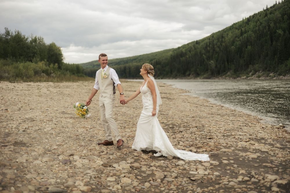 A bride and groom walk hand in hand along a riverbank beneath forested hills.