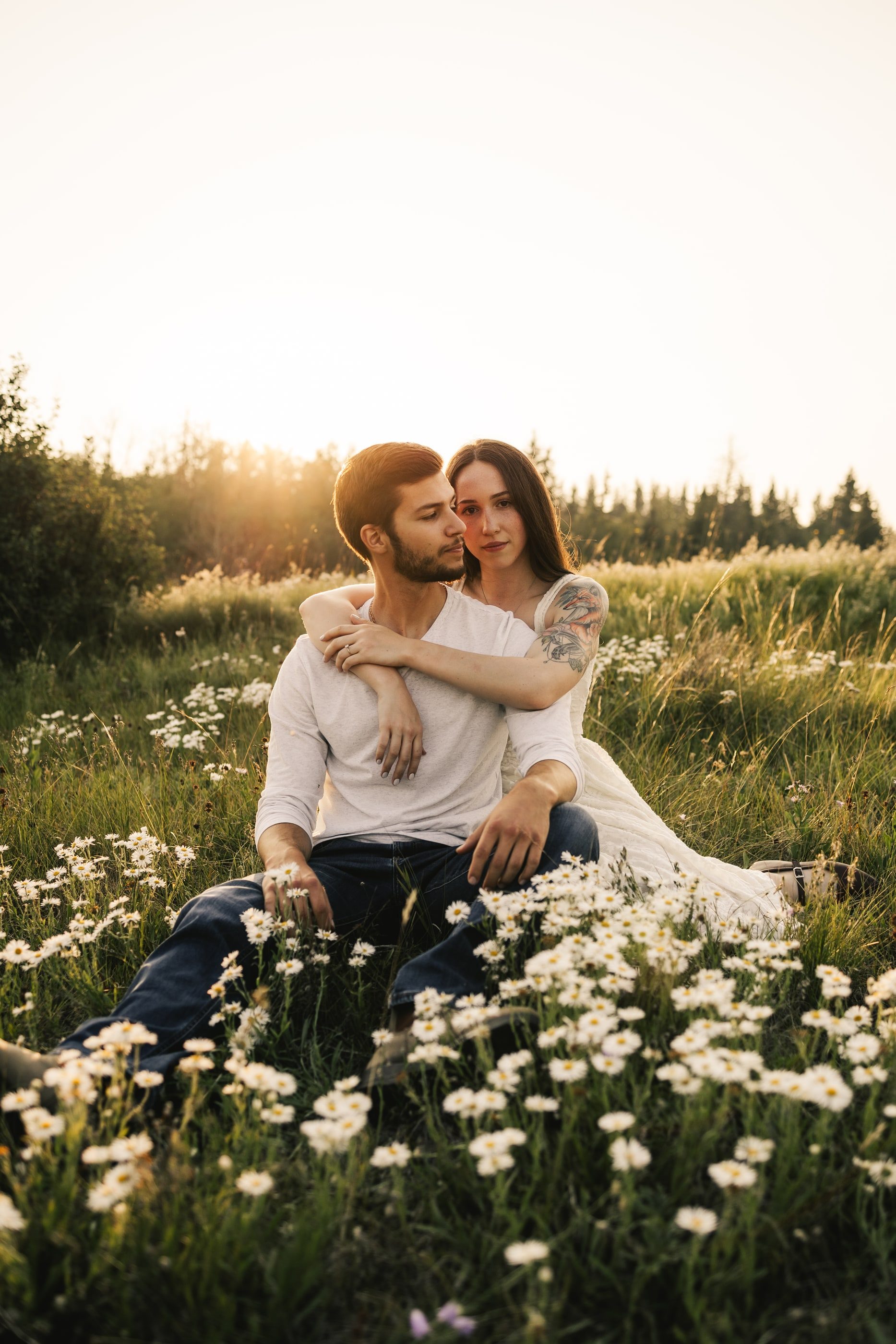 A woman hugs her partner from behind as they sit among white daisies at sunset.