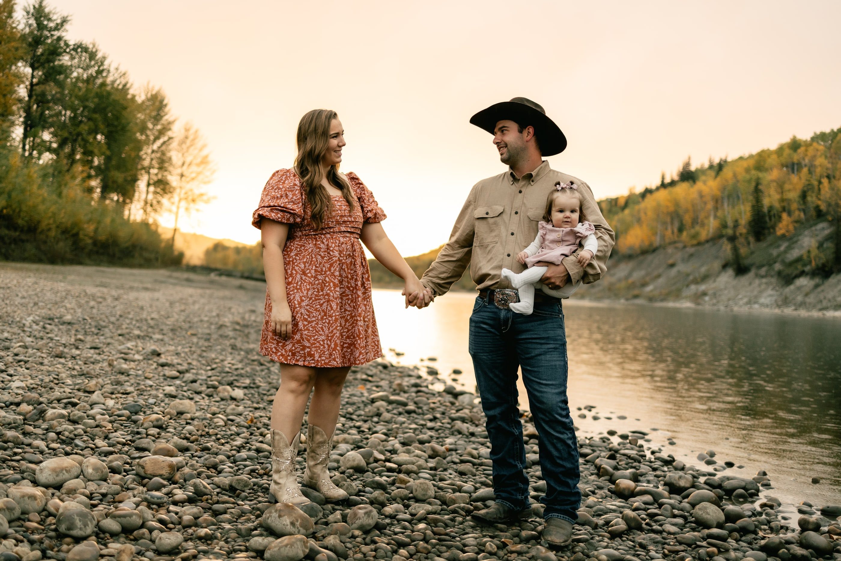 A couple holds hands on a rocky riverbank, the father in a cowboy hat carrying their baby.