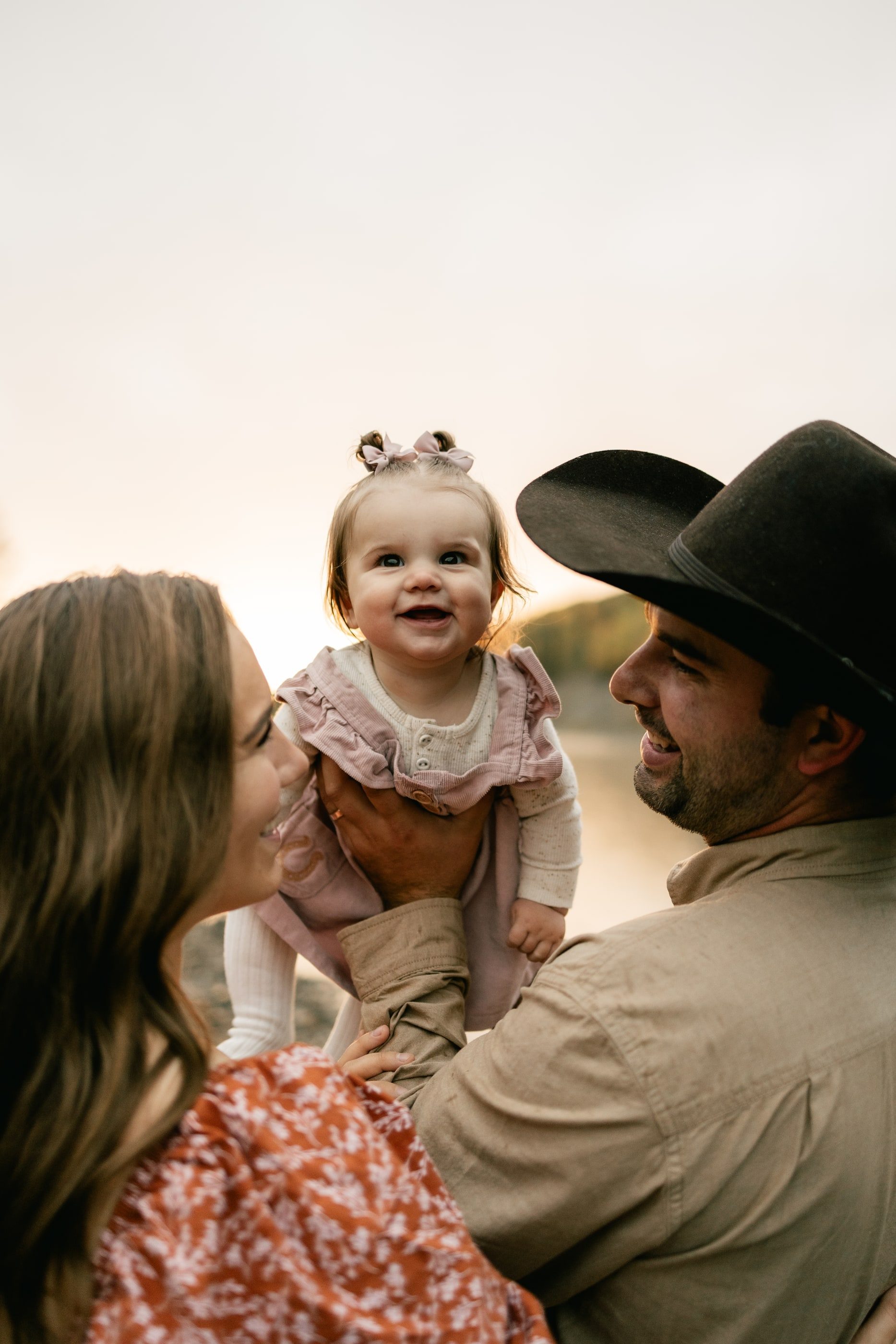 Parents lift their smiling baby girl between them by the river, the father in a cowboy hat.