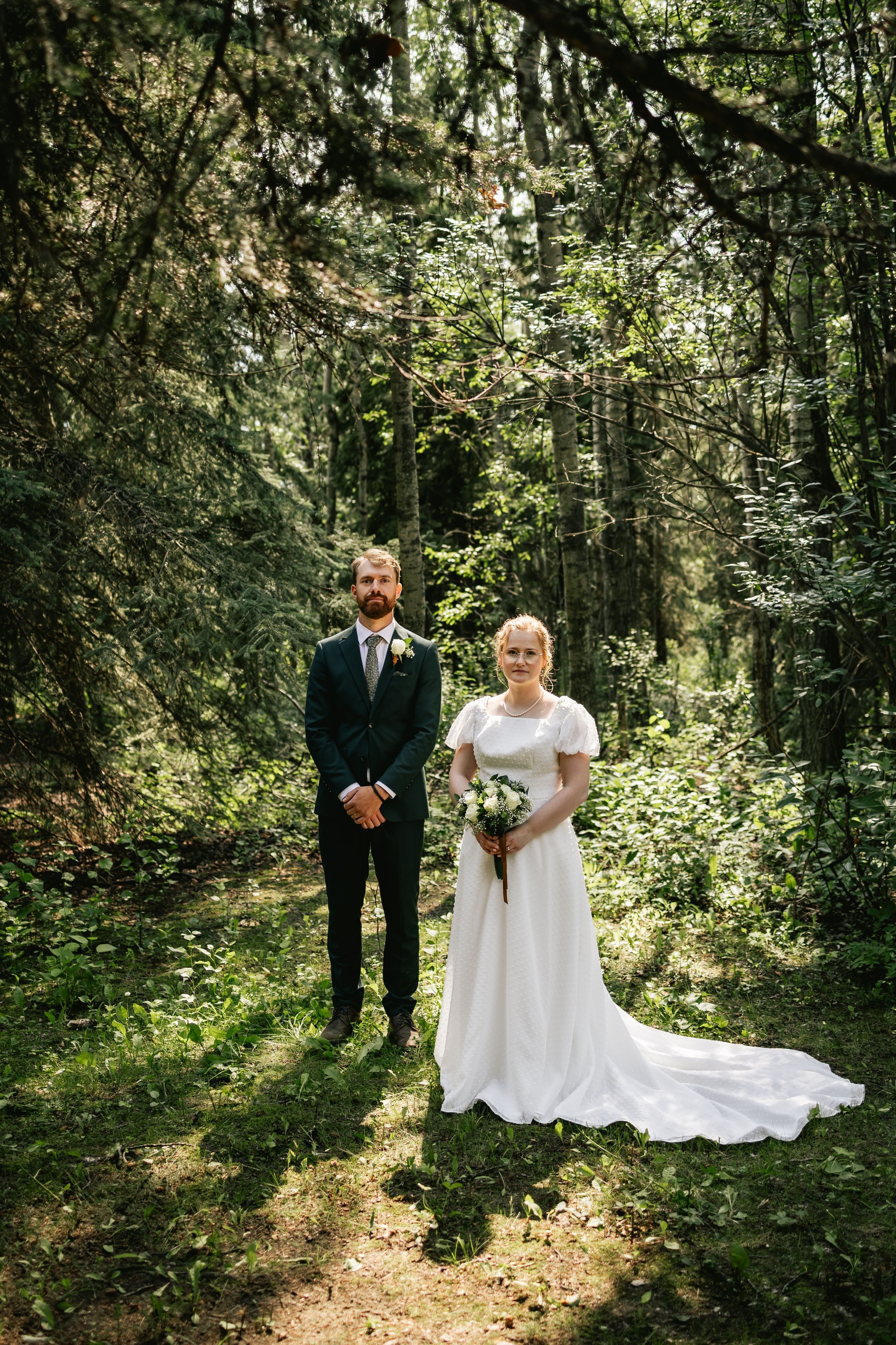 A bride and groom stand side by side in a sunlit clearing in the forest.