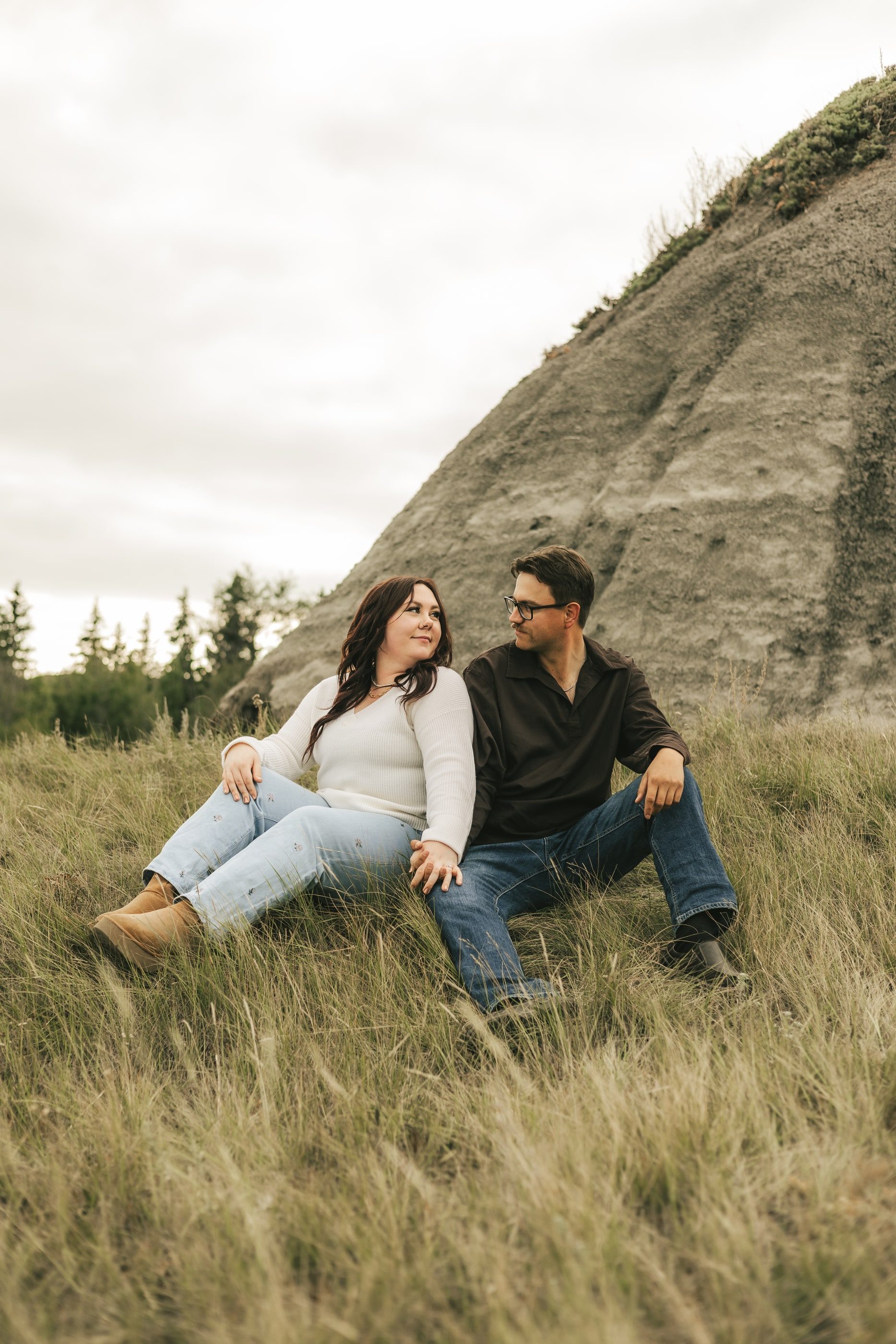 A couple sits together in long grass beneath a steep badlands hill.