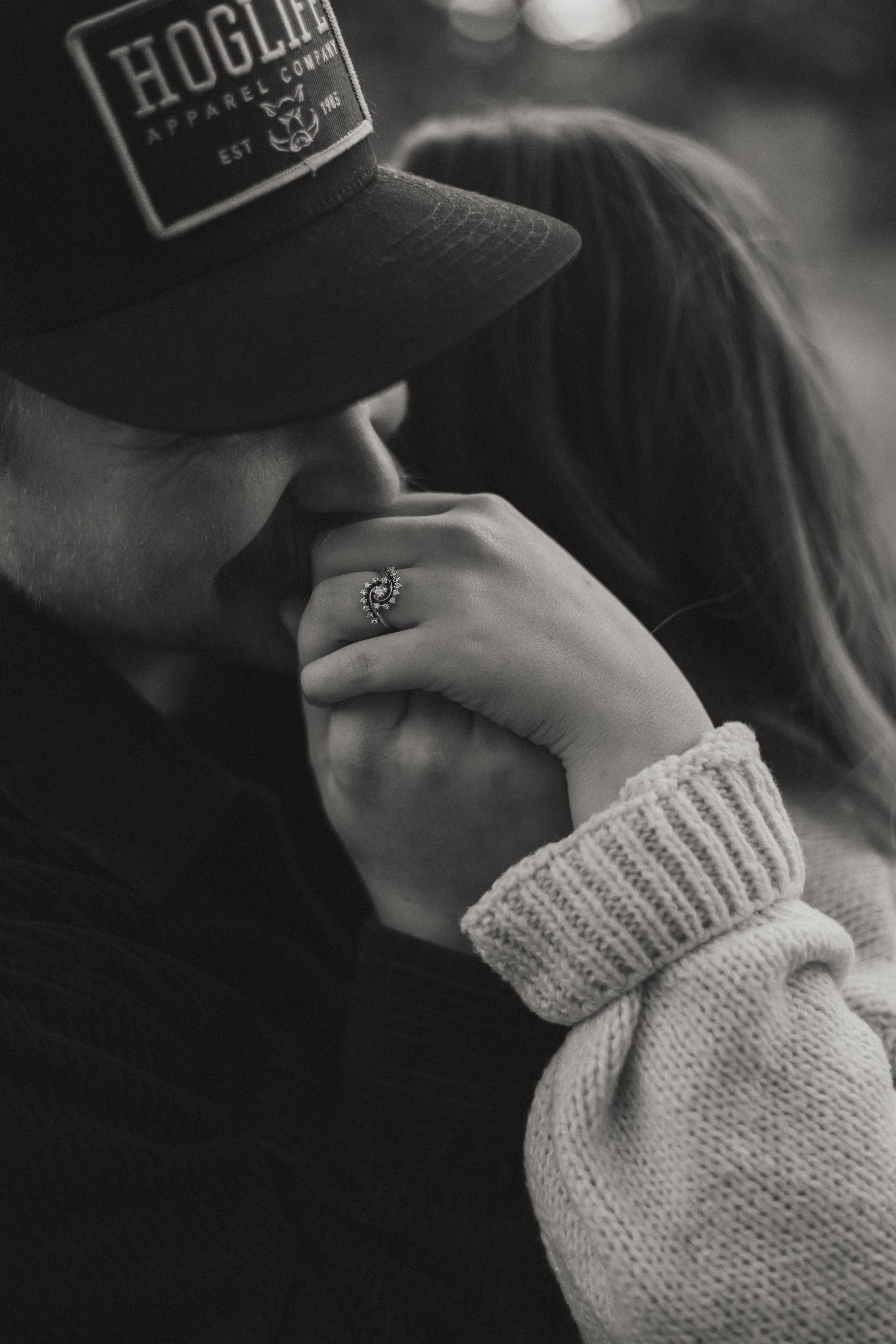 Black-and-white close-up of a man in a cap holding his partner's hand to his lips, her engagement ring showing.