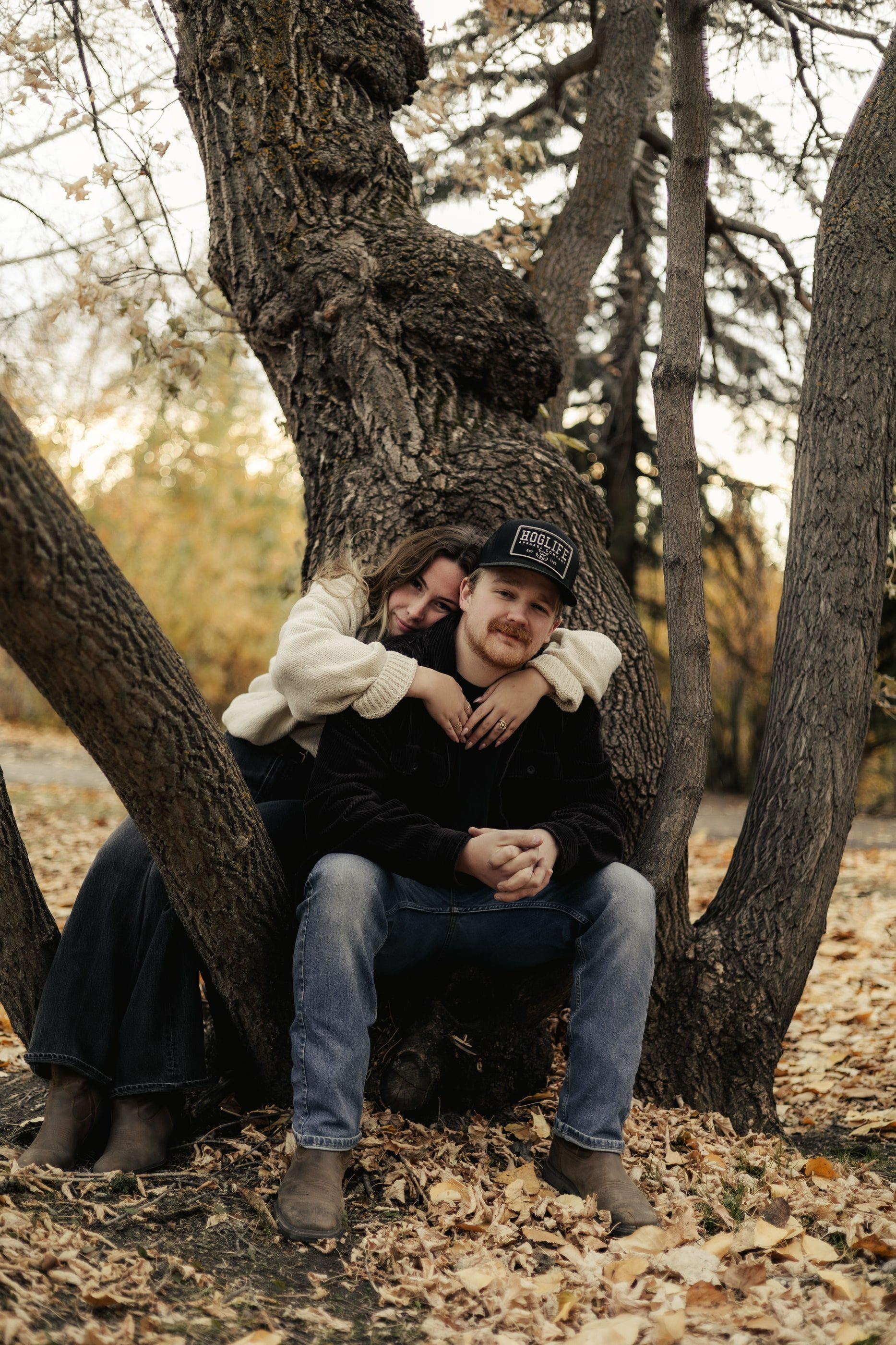 A woman hugs her partner from behind as they sit in the fork of a large tree on autumn leaves.
