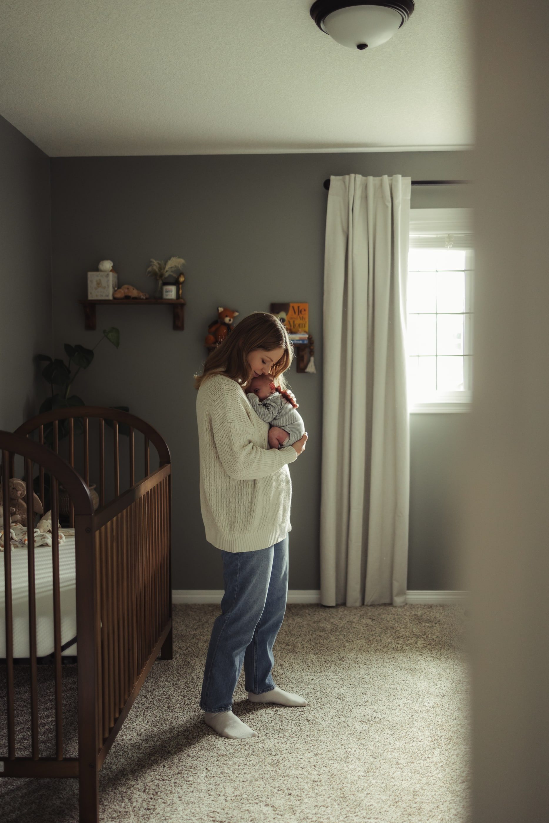A mother cradles her newborn in a softly lit nursery beside a wooden crib.