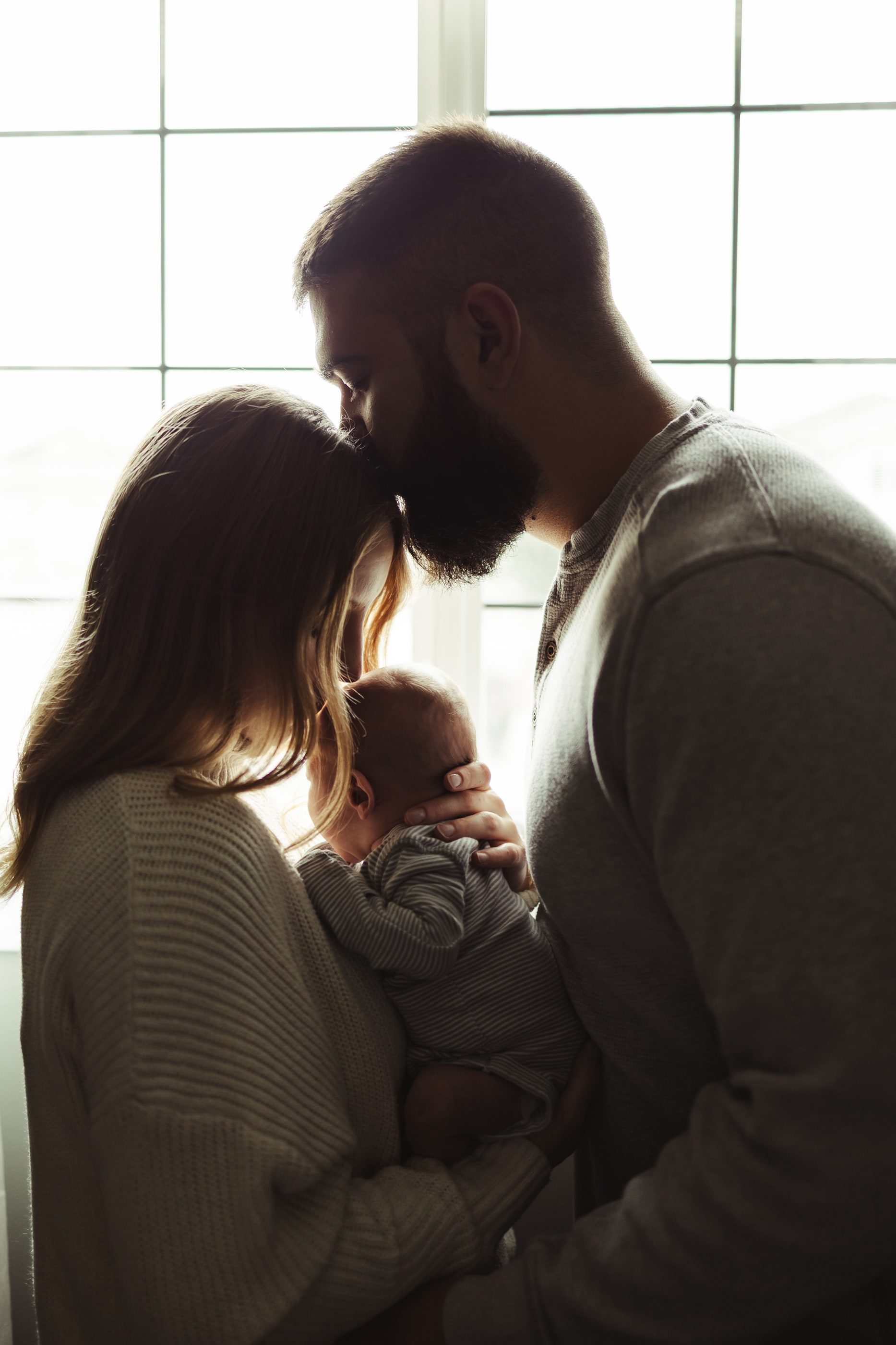 A father kisses the mother's forehead as she holds their newborn by a bright window.