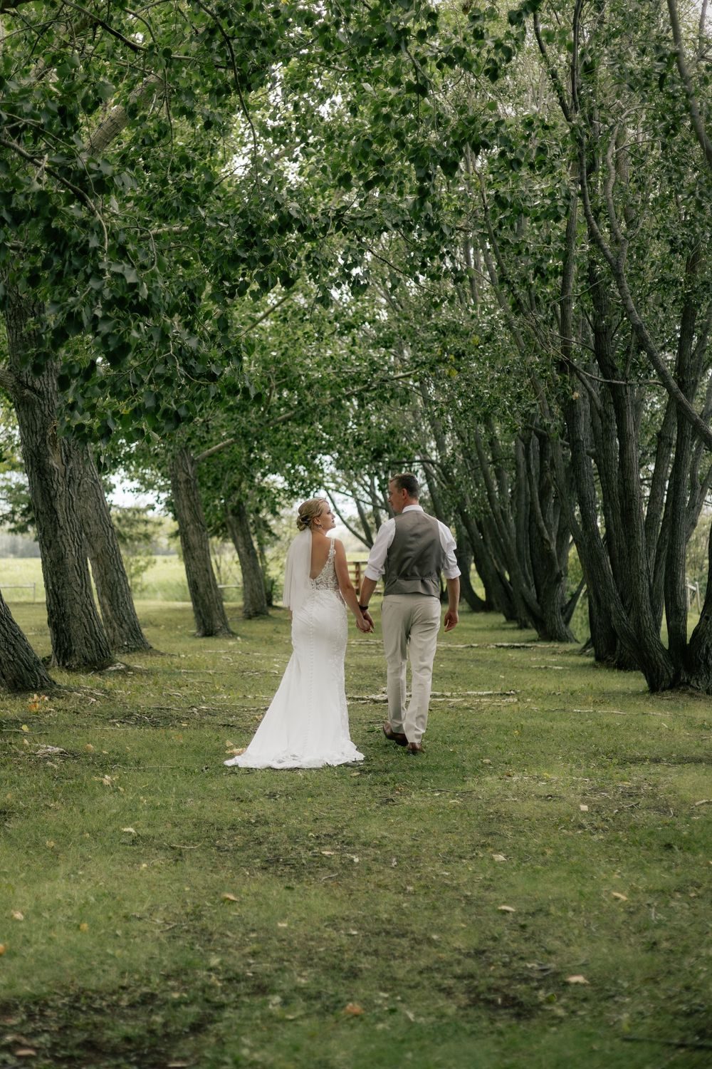 A bride and groom stroll down a long avenue of trees.