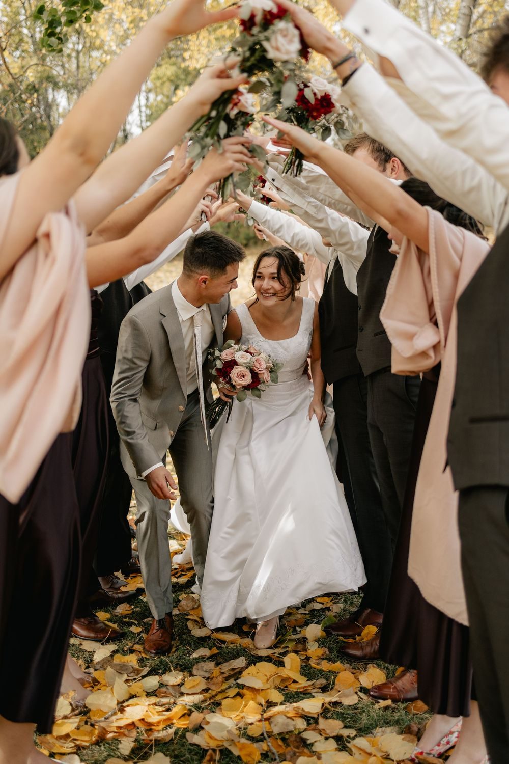 Friends raise their arms into an arch as the bride and groom pass beneath, autumn leaves underfoot.