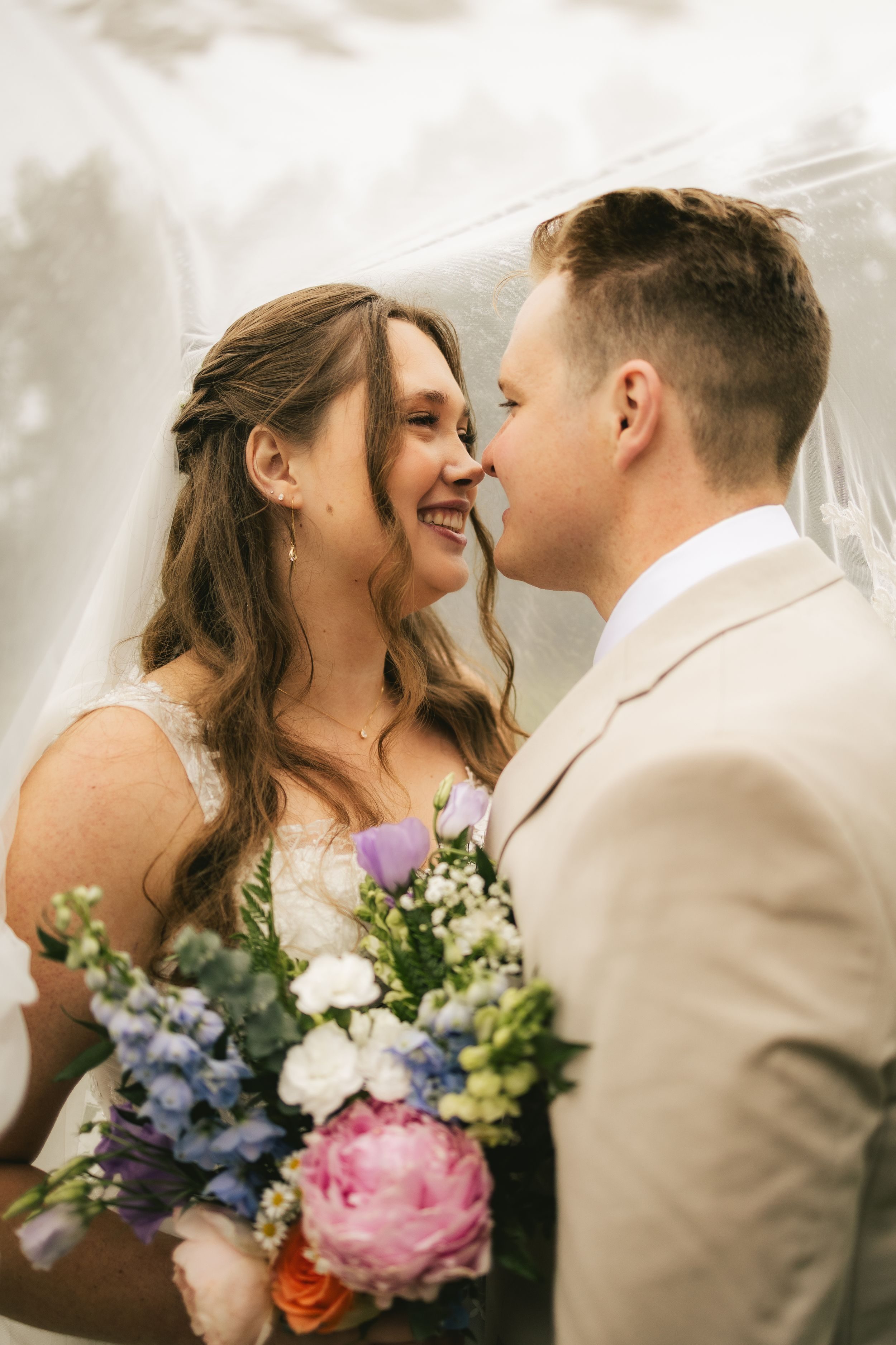 Beneath her veil, a bride and groom touch noses and laugh, holding a bouquet of peonies and delphinium.