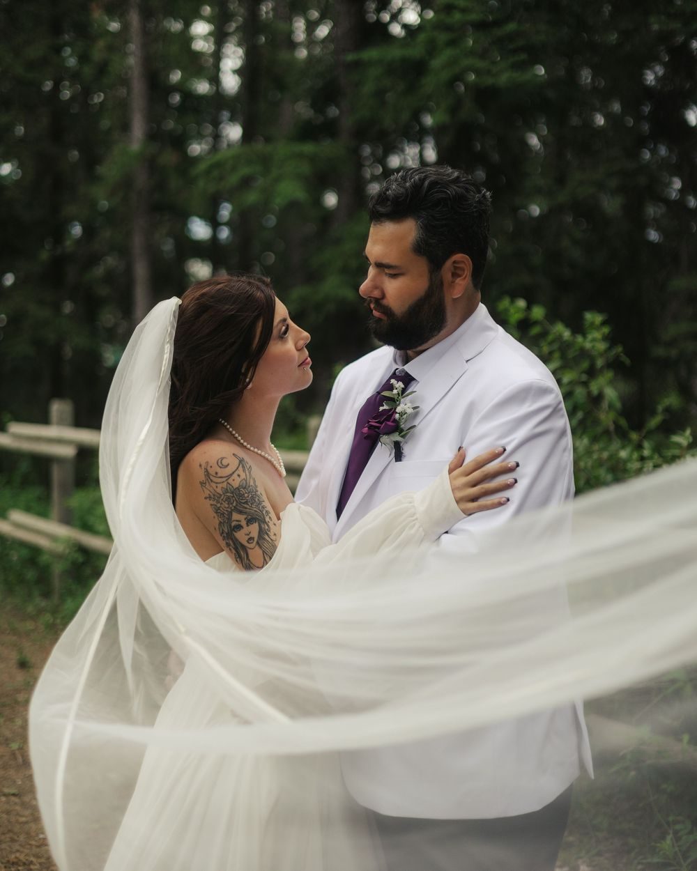 A long veil sweeps across the frame as a bride looks up at her groom in the forest.