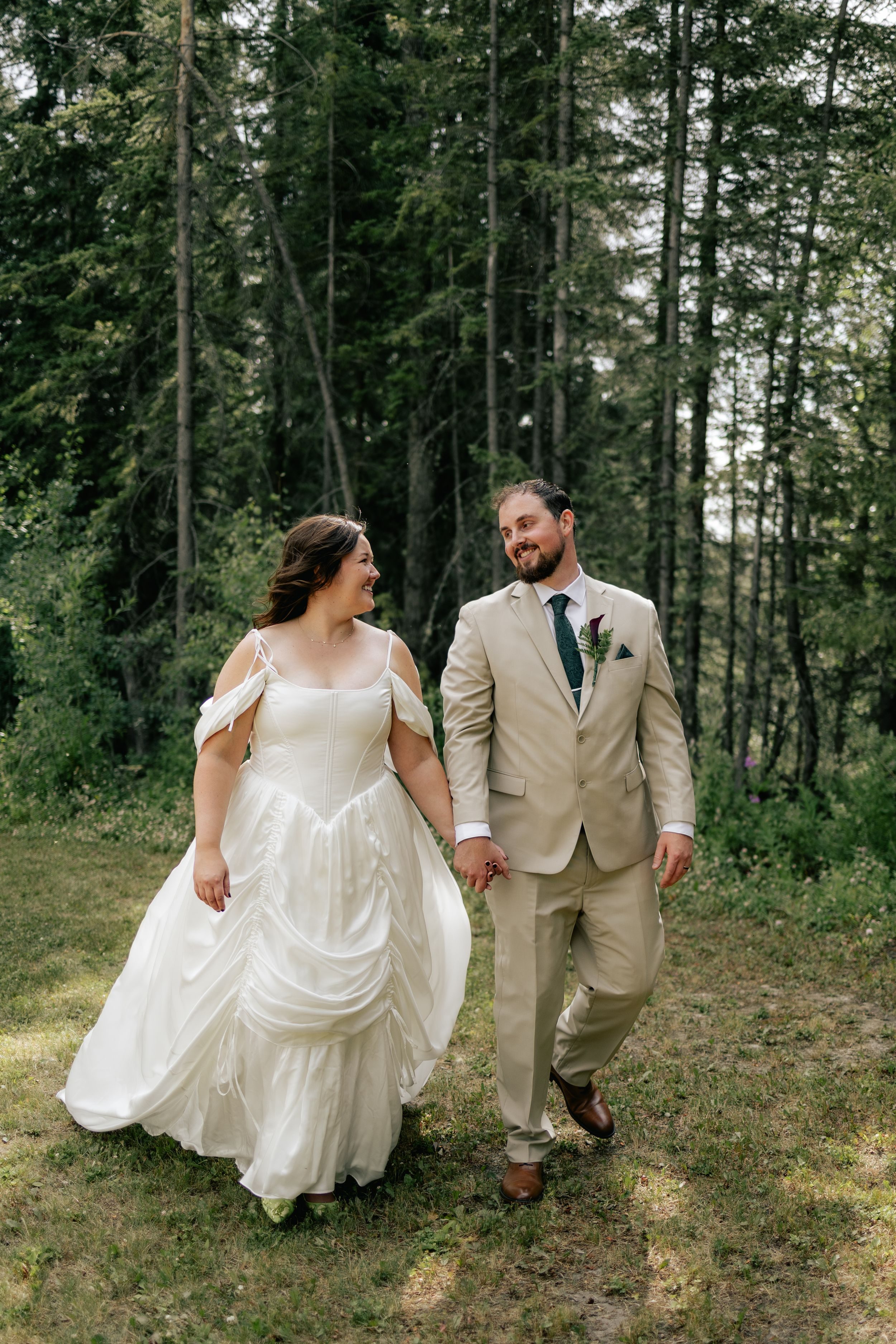 A bride and groom walk hand in hand through a forest clearing, smiling at each other.