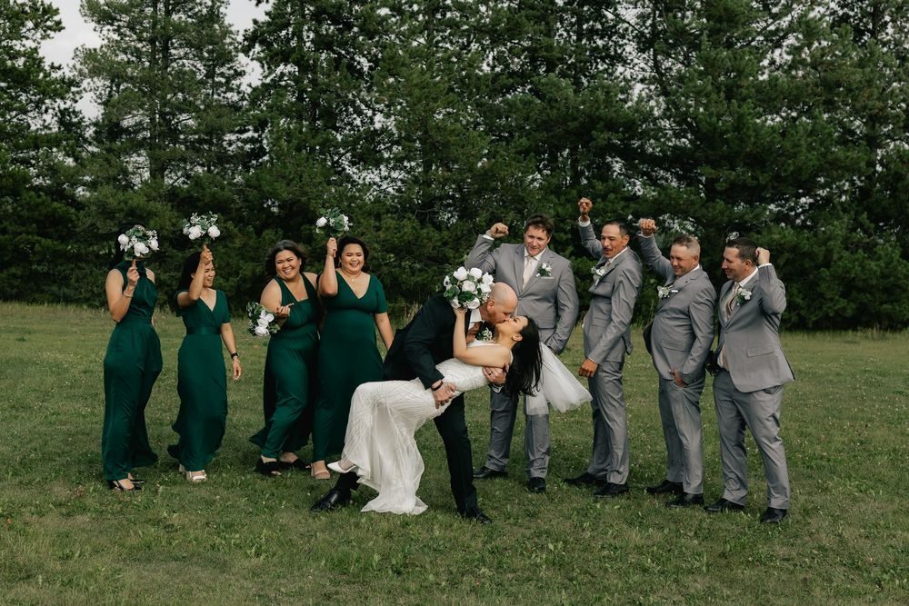 A wedding party in emerald dresses and grey suits cheers as the groom dips the bride on the lawn.