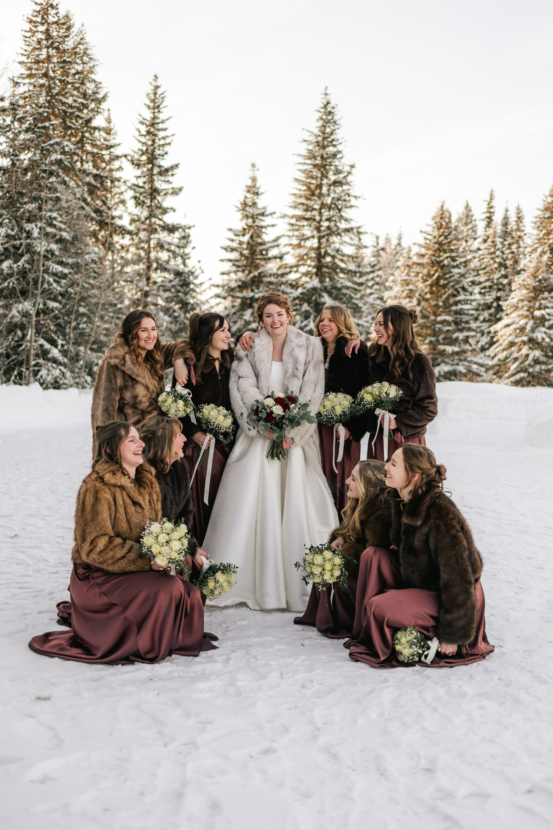 A bride in a white fur wrap stands with her bridesmaids in brown and burgundy among snowy spruce trees.