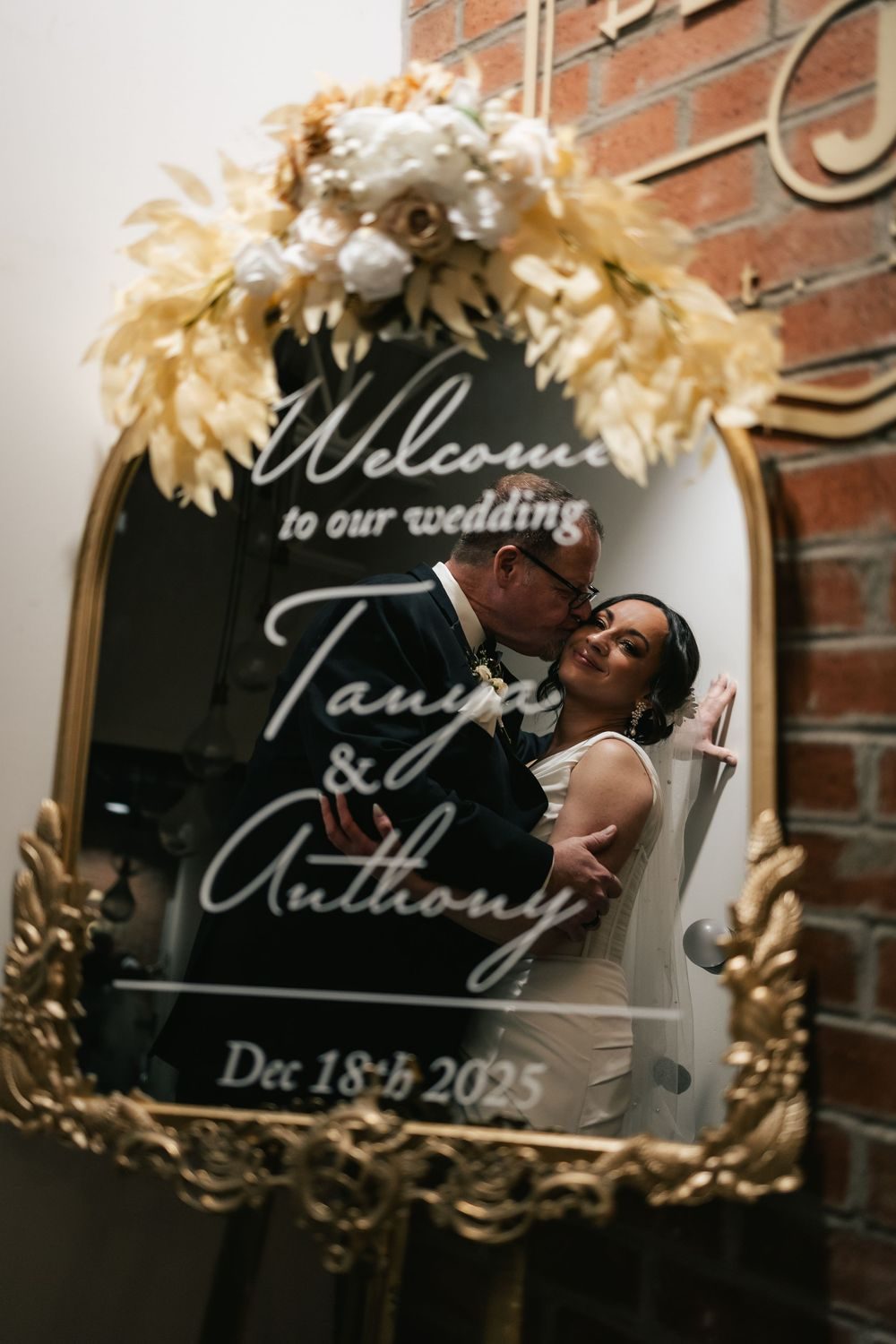 A couple kissing, reflected in a gilded mirror that reads 'Welcome to our wedding'.
