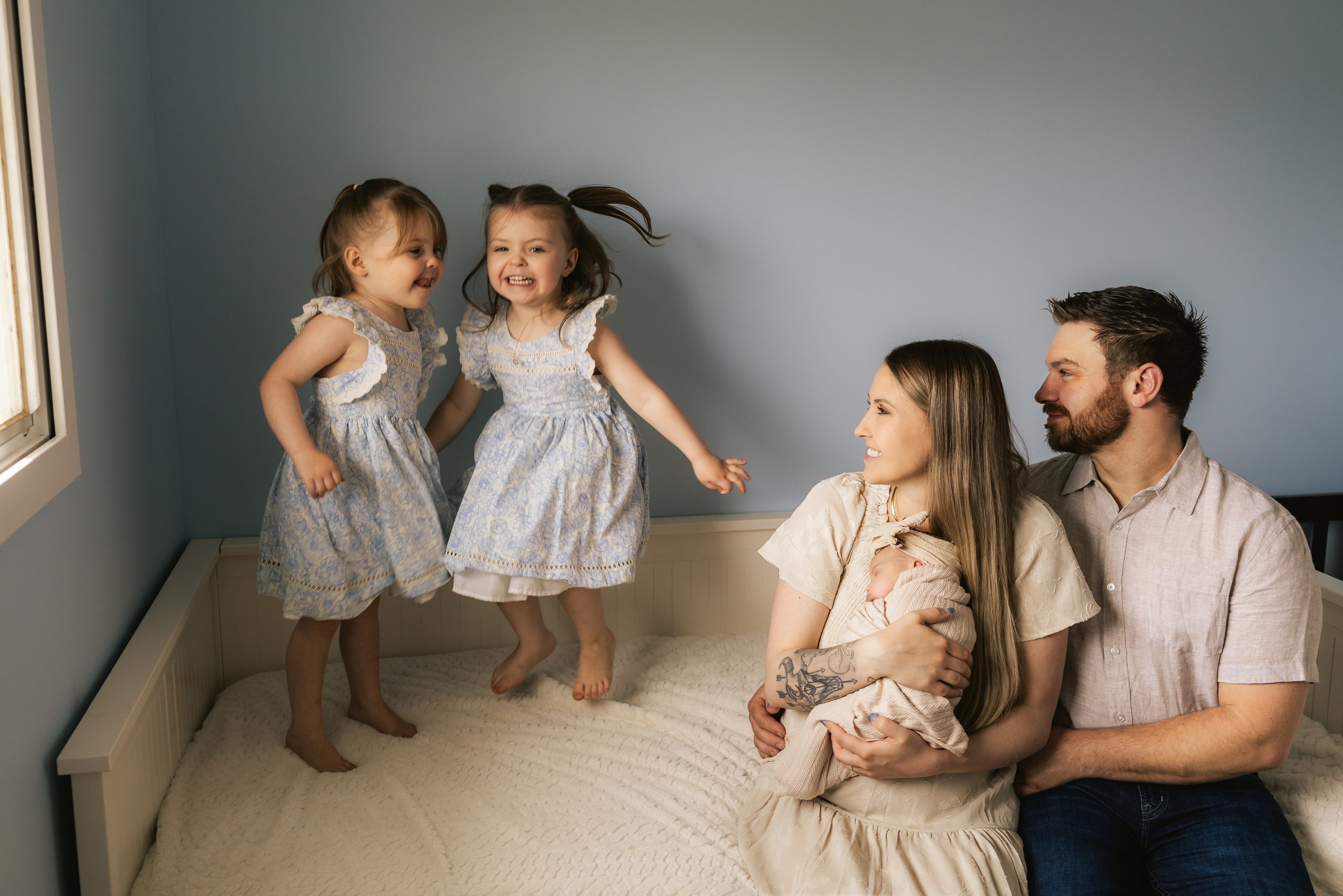 Two little girls jump on a bed as their parents sit beside them holding a newborn.