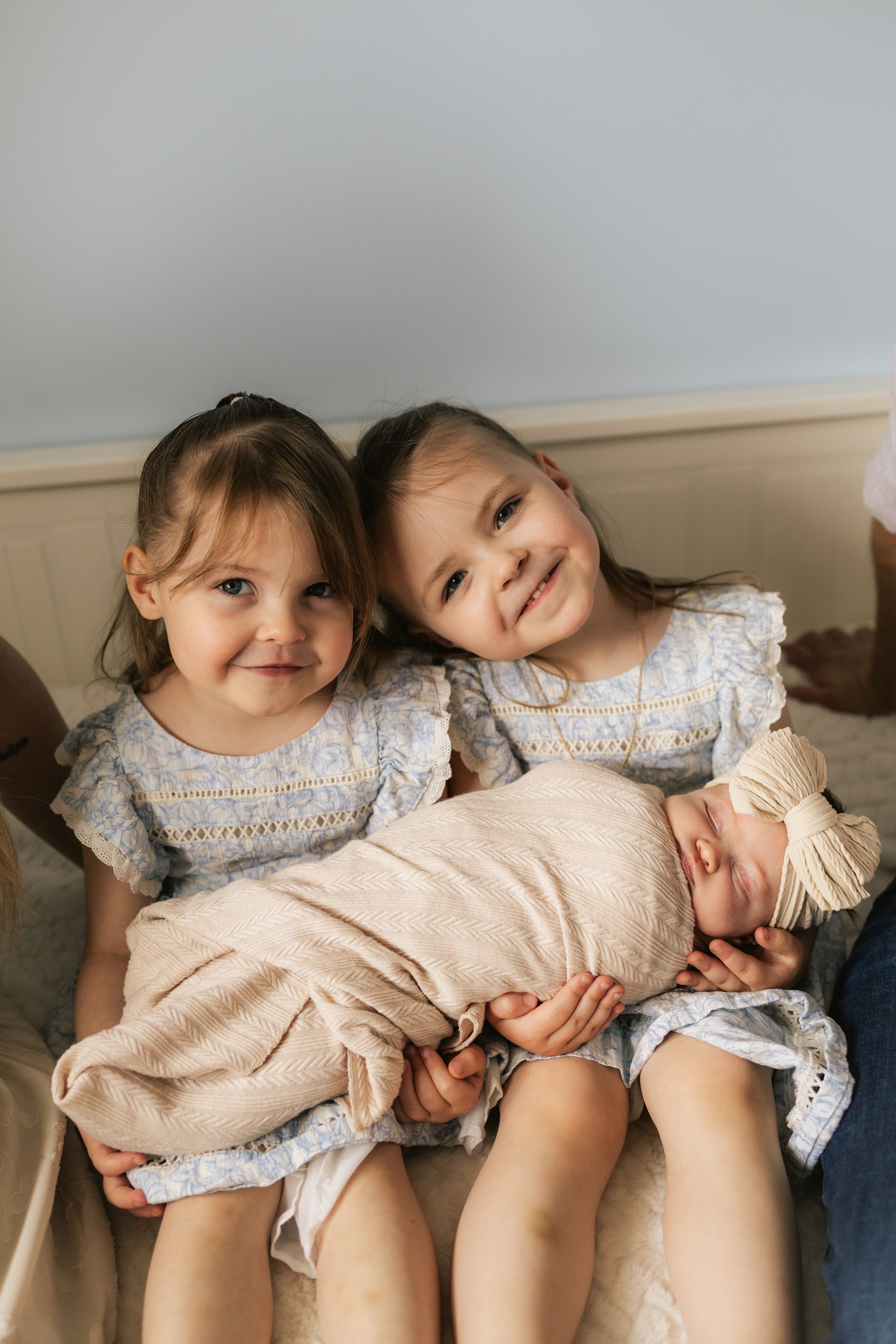 Two little sisters hold their swaddled newborn sibling between them.